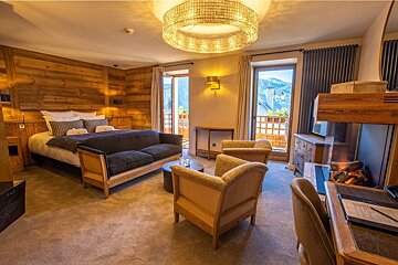 An alpine-style hotel room with wood-paneled walls, a large bed, seating area, desk, electric fireplace, and balcony views.