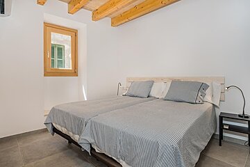 A clean, bright bedroom with twin beds, striped bedding, wooden ceiling beams, and a wooden-framed window with green shutters. Minimalist design.