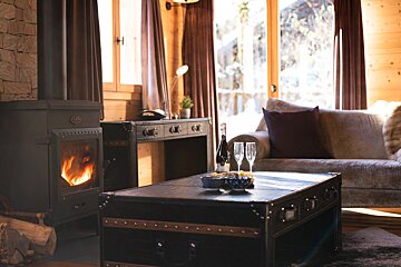 A cozy cabin living room with a lit fireplace, champagne on a trunk coffee table, and a sofa, overlooking a bright winter scene.
