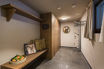 A modern rustic entryway features a wooden bench, shoe dryer, wood cabinets, tiled floor, and shelf. Pillows, a fruit bowl, and a mirror decorate the space.