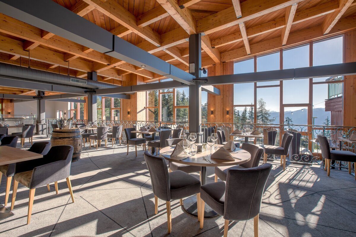 An elegant, sunlit restaurant with wooden beamed ceilings, expansive windows showcasing mountain vistas, and tables set for dining.