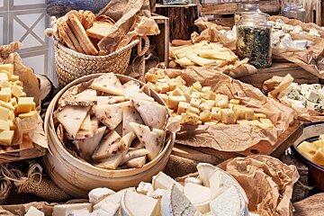 A rustic buffet spread with an abundance of various cheeses (cubed, sliced, wedges, blue) and bread, displayed on kraft paper and in baskets.
