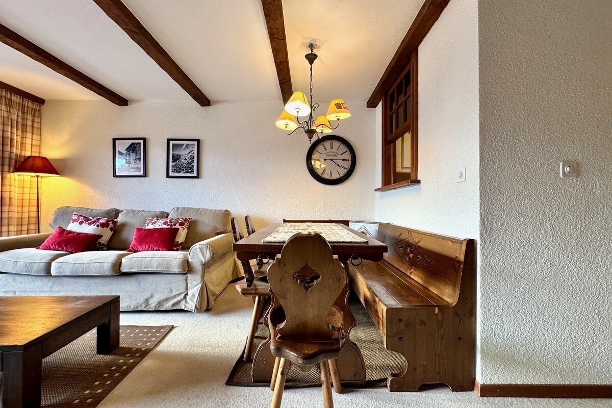 A cozy, rustic living/dining room with exposed ceiling beams, a comfy beige sofa with red pillows, and wooden dining furniture featuring a bench and wall clock.