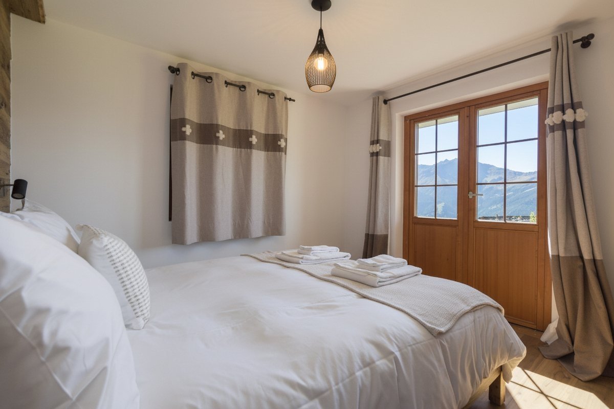Cozy bedroom with white bedding, patterned curtains, and double doors opening to a stunning mountain view. A warm light fixture hangs from the ceiling.