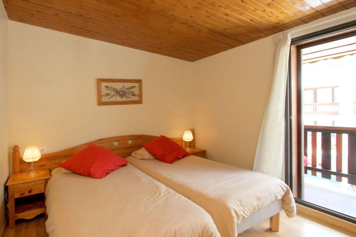 A cozy bedroom with two single beds, red pillows, wooden furniture, and a textured wooden ceiling. A large window opens to a balcony.