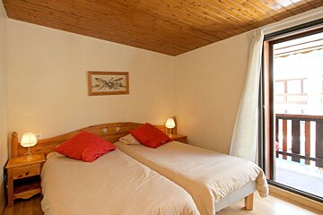 A cozy bedroom with two single beds, red pillows, wooden furniture, and a textured wooden ceiling. A large window opens to a balcony.