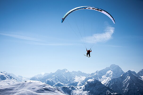 A person skispeedflying with a blue paraglider, soaring over vast snow-capped mountains against a clear blue sky.