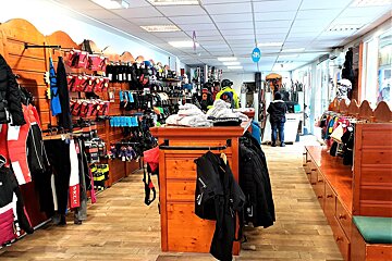 A brightly lit ski and winter sports shop filled with colorful apparel, gloves, and gear on wooden racks and displays, with customers browsing.