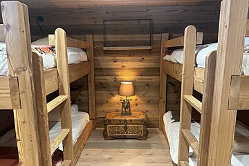 Rustic bunk room with two wooden bunk beds flanking a central chest with a lamp and a shelf. Wood paneling covers walls and ceiling.