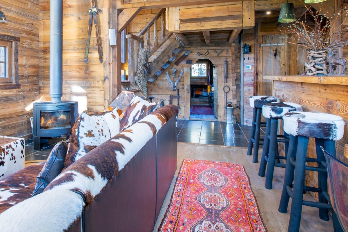 A cozy, rustic chalet interior features abundant wood, a warm fireplace, cowhide-covered seating, and a bar with matching stools.
