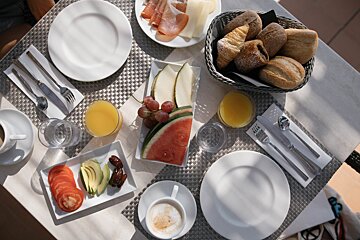 A bright, varied breakfast spread with fruit, bread, ham, cheese, juice, and coffee on an outdoor table.
