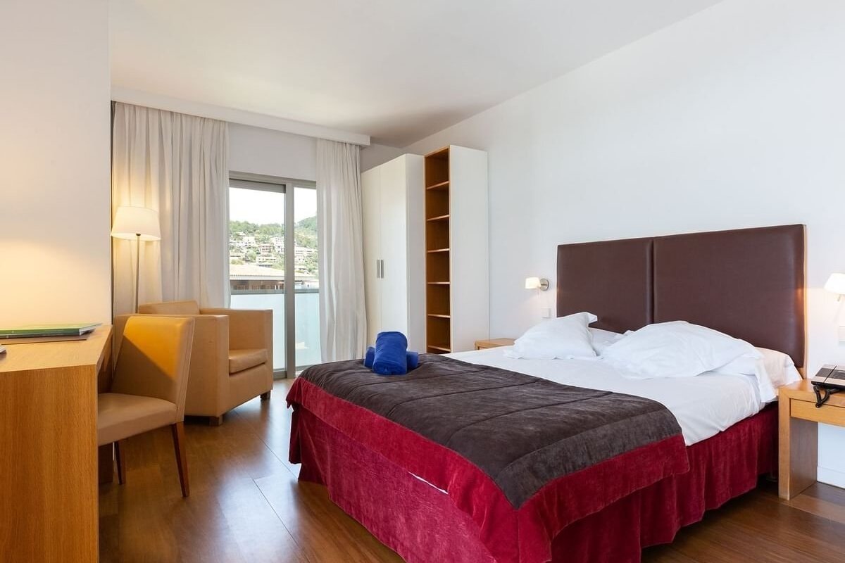 A bright hotel bedroom with a large bed, red and brown covers, wooden floor, and a balcony offering a scenic view of a hillside town.