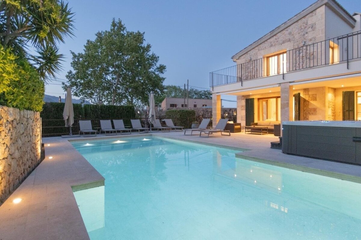 Twilight scene of a stone villa with a brightly lit swimming pool, jacuzzi, and patio. Loungers line the pool.