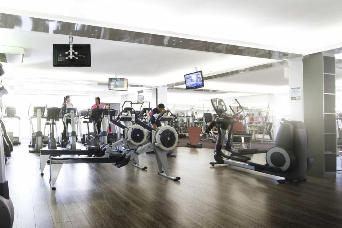 A bright, spacious gym with several people exercising on various cardio machines like treadmills, rowing machines, and ellipticals. TVs hang from the ceiling.