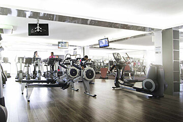 A bright, spacious gym with several people exercising on various cardio machines like treadmills, rowing machines, and ellipticals. TVs hang from the ceiling.