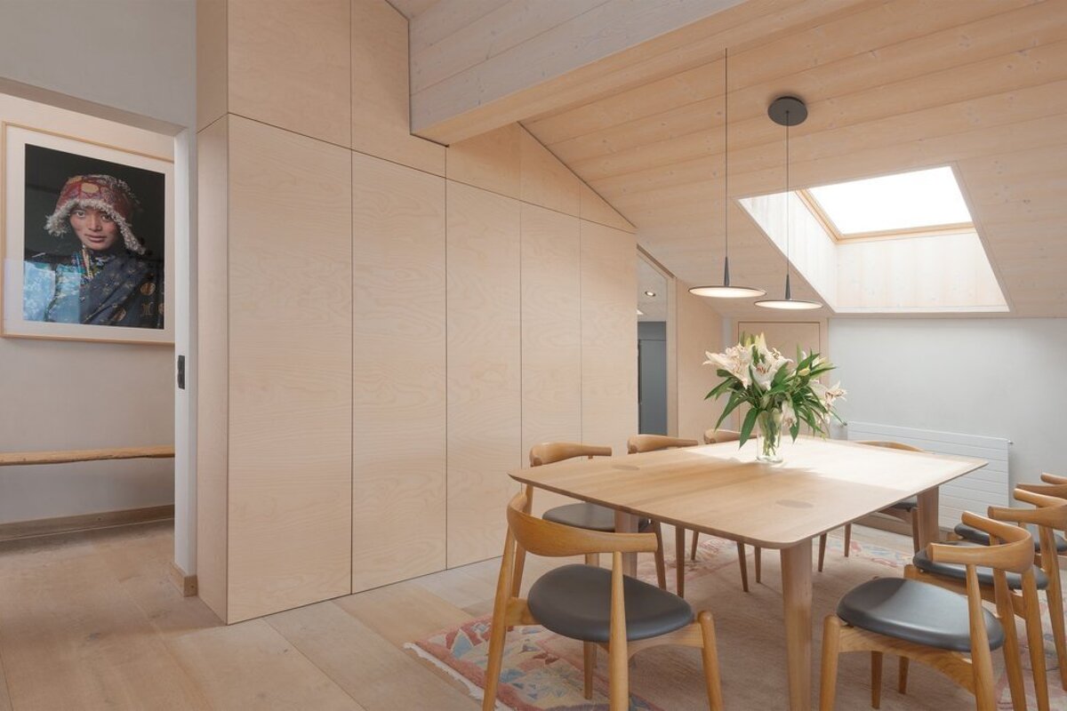Bright, modern dining area with light wood table, chairs, and extensive cabinetry. A skylight and pendant lights illuminate the space, with flowers on the table and framed artwork visible.