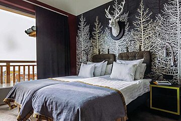 A rustic-modern bedroom with twin beds, dark forest wallpaper featuring a deer head, and a balcony with outdoor views.