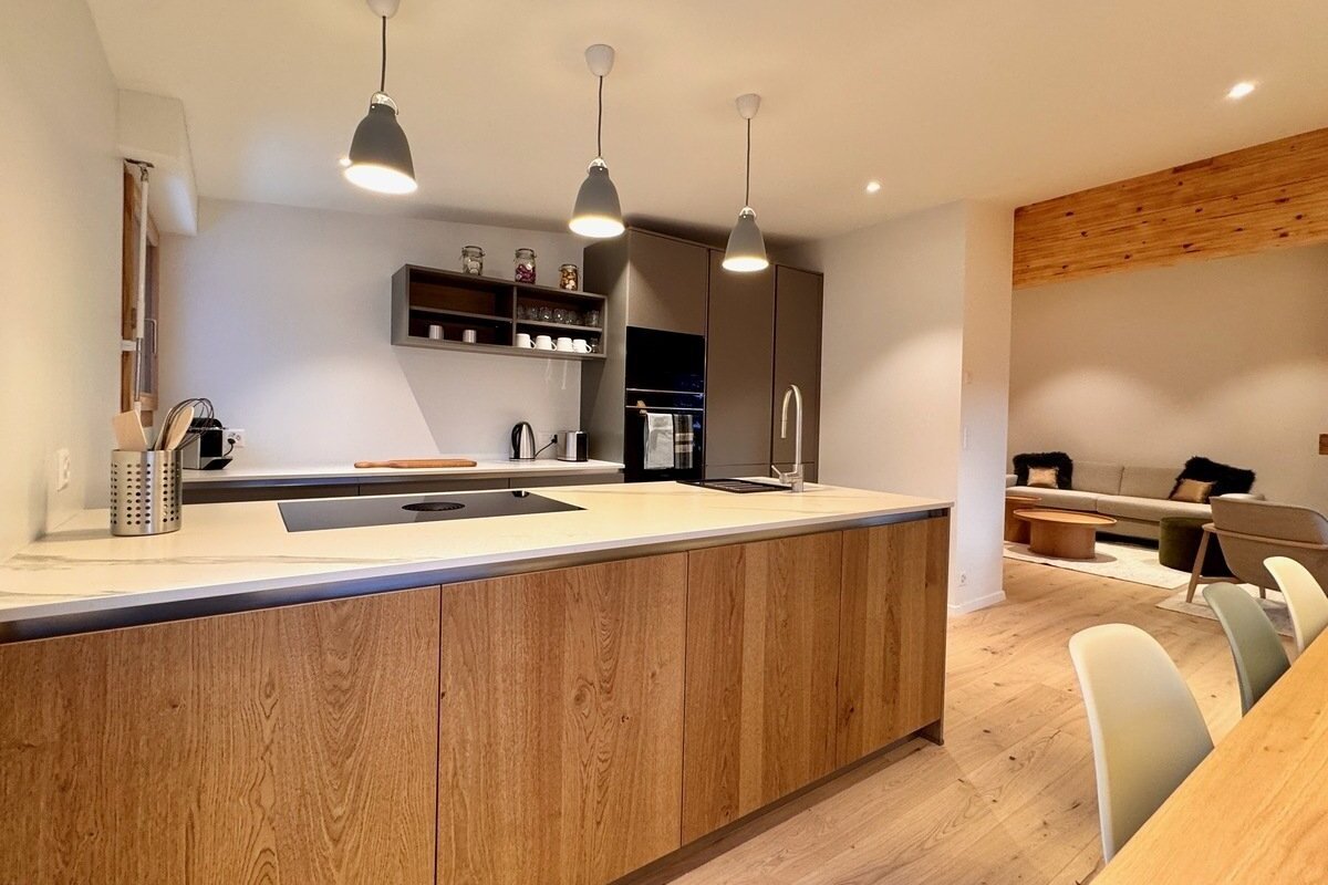A modern open-plan kitchen with a large wooden island, light countertops, and grey built-in appliances, flowing into a comfortable living room.