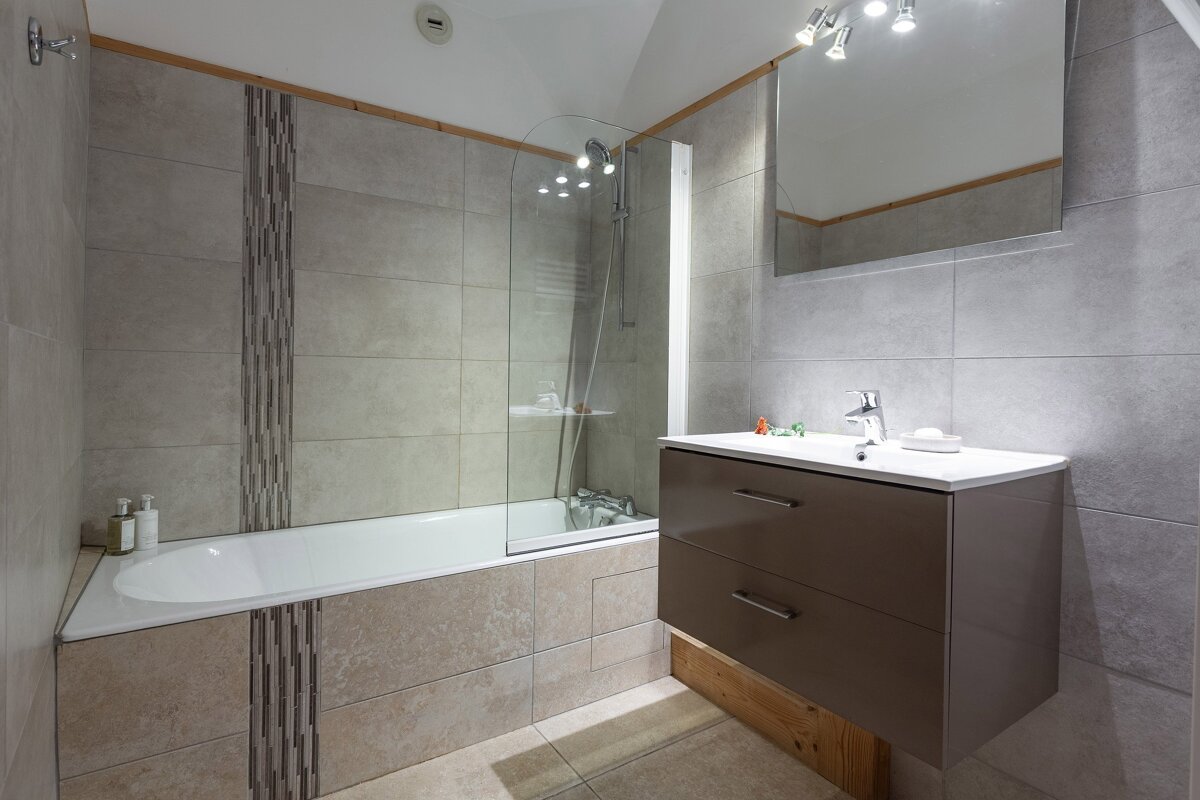 A modern bathroom with grey tiled walls, a bathtub/shower combo, and a floating brown vanity with a sink and mirror. Wooden accents are visible.
