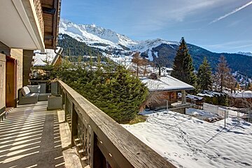 A sunny balcony overlooks a stunning snowy mountain landscape with pine forests and chalets, all under a clear blue sky.