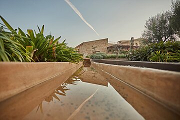 A low-angle view shows a water channel reflecting stone buildings, green plants, and a sky with a distinct contrail.