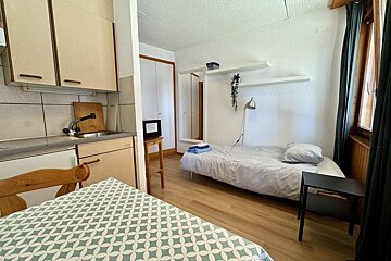 Cozy studio apartment with a kitchenette, single bed, dining table, and window. White walls, wooden floor, and simple furniture.