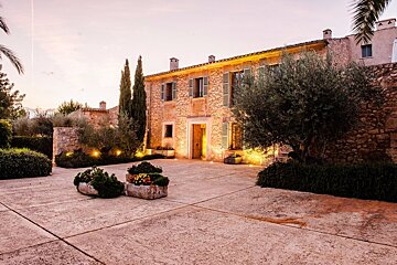 Mediterranean stone villa at dusk, warmly lit, with olive trees, cypress, and a paved courtyard.