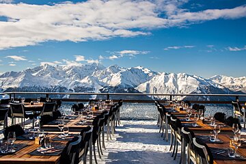 A snowy outdoor restaurant terrace with set tables, offering breathtaking panoramic views of majestic snow-capped mountains under a bright blue sky.
