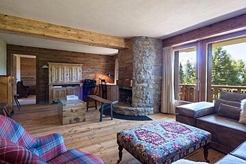 Rustic, wood-paneled living room with a stone fireplace, cozy furnishings, and large windows offering a scenic mountain and forest view.