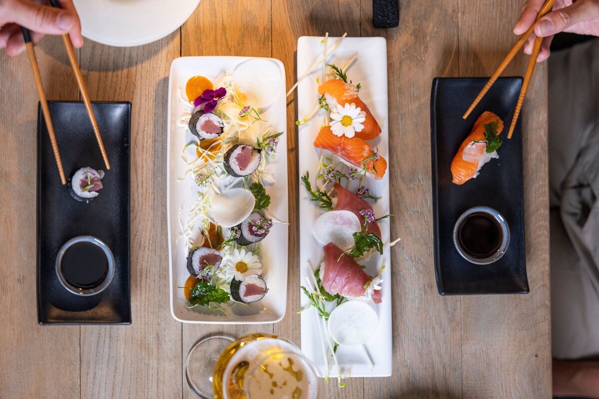 An overhead view of beautifully arranged sushi and sashimi platters. Two pairs of chopsticks pick up pieces, with a beer glass on a wooden table.