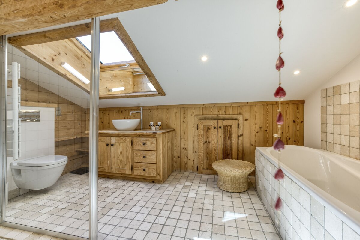 Rustic attic bathroom with wooden walls, ceiling beams, and skylights. Features a white tub, shower, toilet, vanity, and light tiled floor.