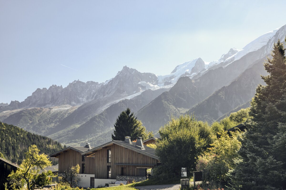 Wooden alpine chalets amidst lush trees, with towering, sunlit, snow-capped mountains forming a majestic backdrop under a clear sky.