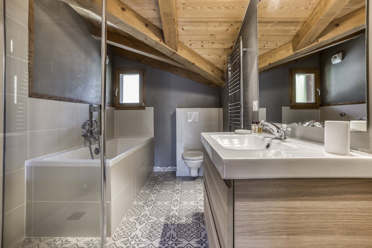 A modern attic bathroom with a sloped wooden ceiling, grey walls, and patterned floor. Features a bathtub, toilet, vanity sink, window, and glass shower screen.