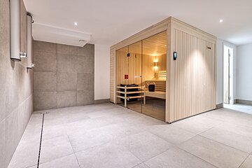 A modern spa room featuring a glass-fronted wooden sauna with interior lighting, adjacent to a tiled shower area and light-colored tiled floors.