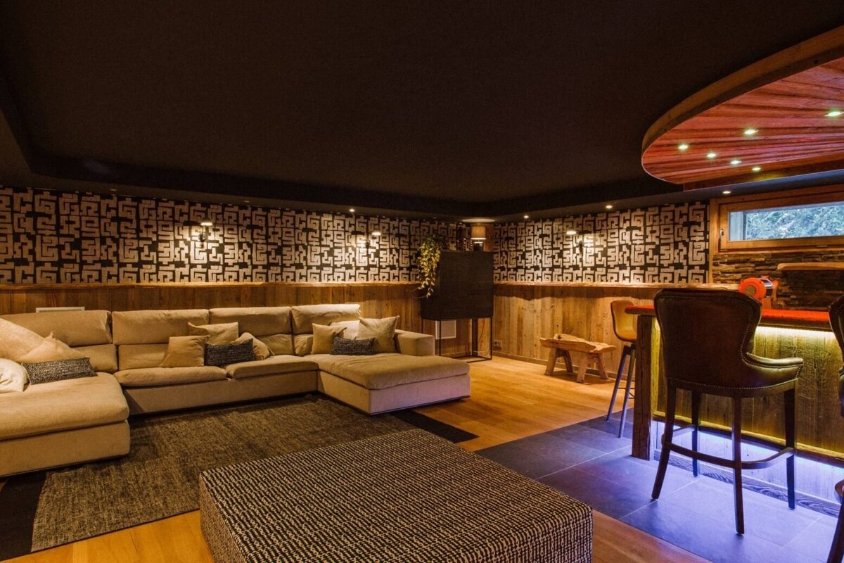 A modern, rustic-themed living space with a large beige sectional sofa, patterned accent wall, wooden wainscoting, and a bar with underglow lighting.