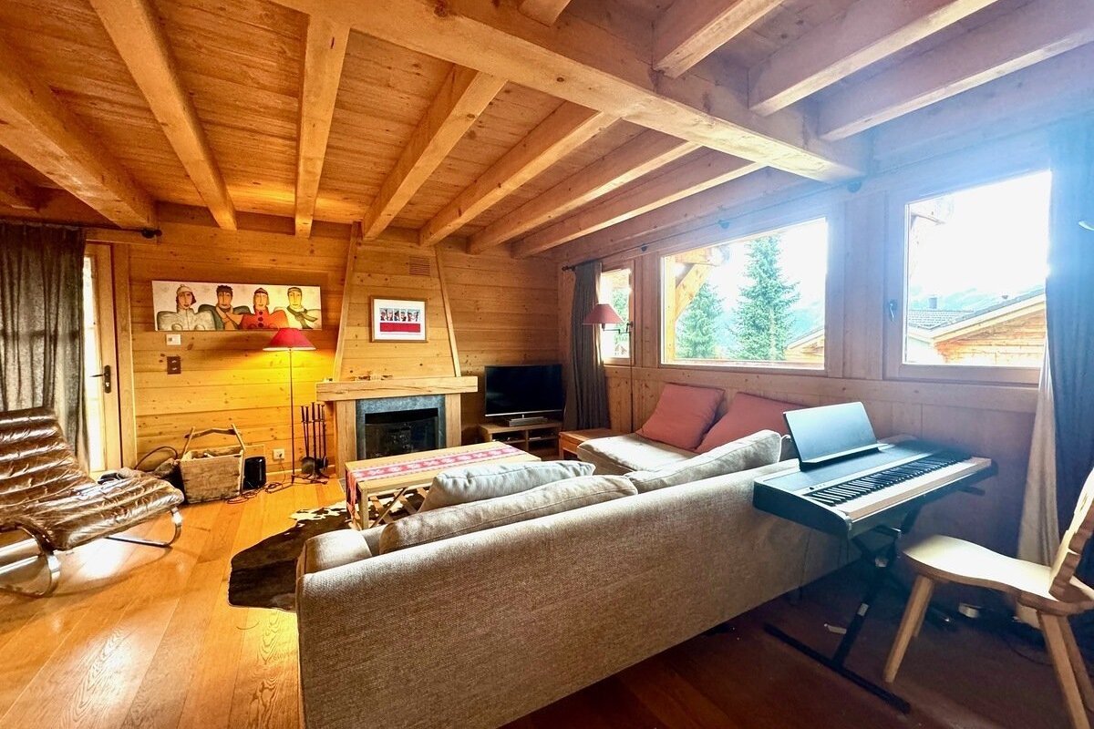 A cozy, wood-paneled chalet living room with a fireplace, sofa, and electronic piano. Large windows offer views of pine trees and neighboring chalets.