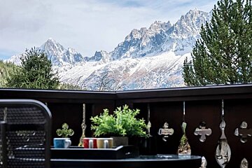 A balcony offers a stunning view of majestic snow-capped mountains and green trees. A table with a plant and colorful cups sits in the foreground.
