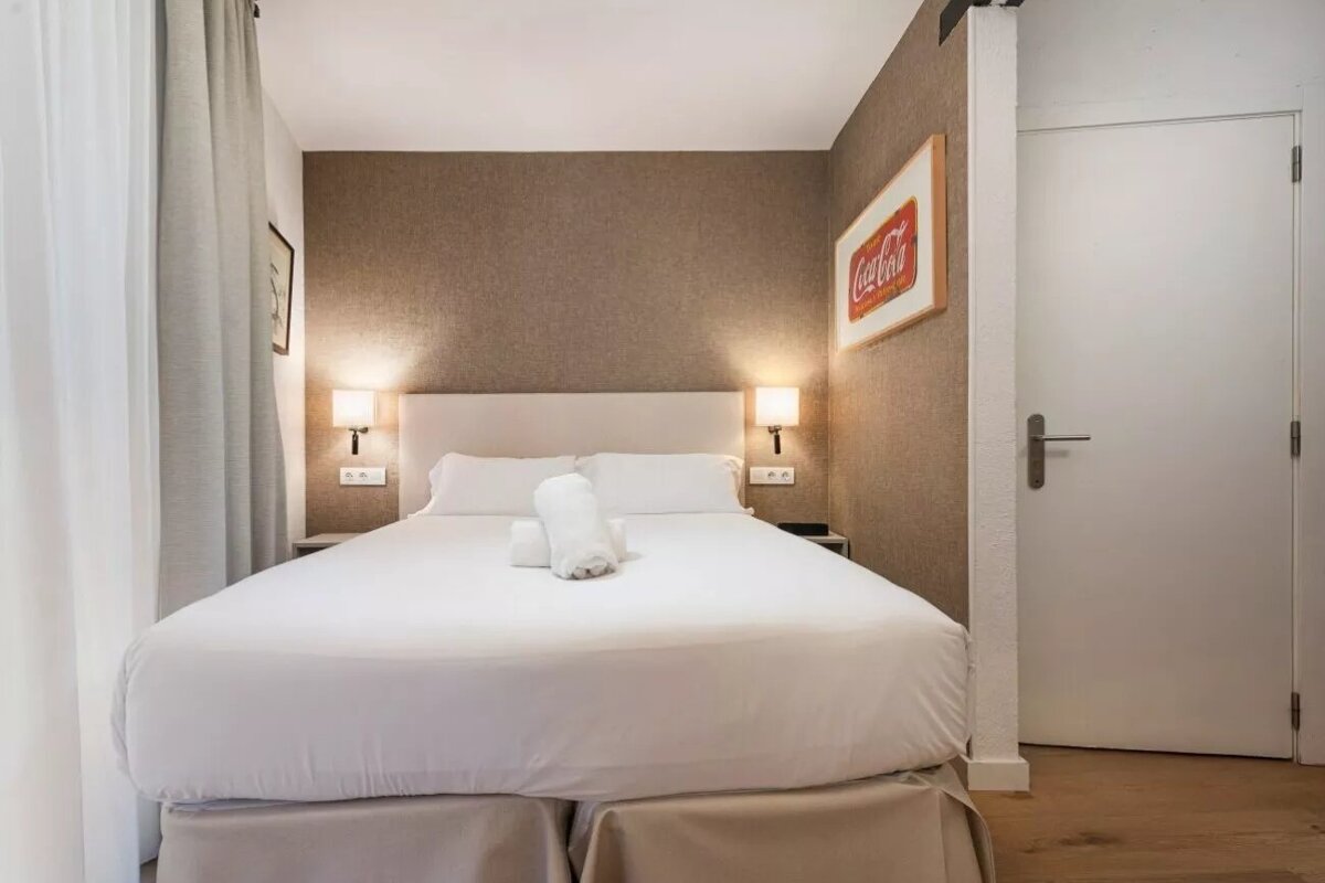 A hotel-style bedroom features a neatly made bed with white linens, two rolled towels, bedside lamps, and a Coca-Cola sign on a brown wall.
