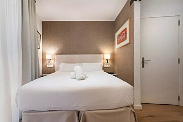 A hotel-style bedroom features a neatly made bed with white linens, two rolled towels, bedside lamps, and a Coca-Cola sign on a brown wall.