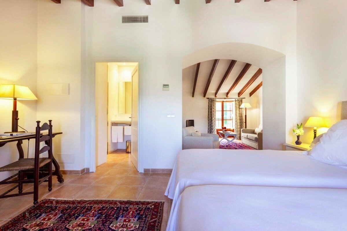 A light-filled hotel suite with a bedroom, desk, bathroom, and an arched opening to a living area with sofas, wooden beams, and tiled floors.