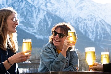 Friends laugh and drink beer on a sunny mountain patio.