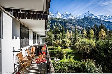 A sunny balcony with chairs and flowers overlooks a green garden and forest, with majestic snow-capped mountains under a clear blue sky.