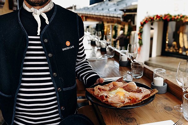 A person in a Breizh Cafe vest and striped shirt holds a savory crepe with ham and egg, dining al fresco at a festive outdoor cafe.