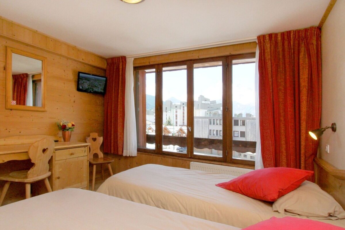 A cozy, wood-paneled bedroom with two twin beds, red curtains, a desk, and a large window looking out onto buildings and mountains.