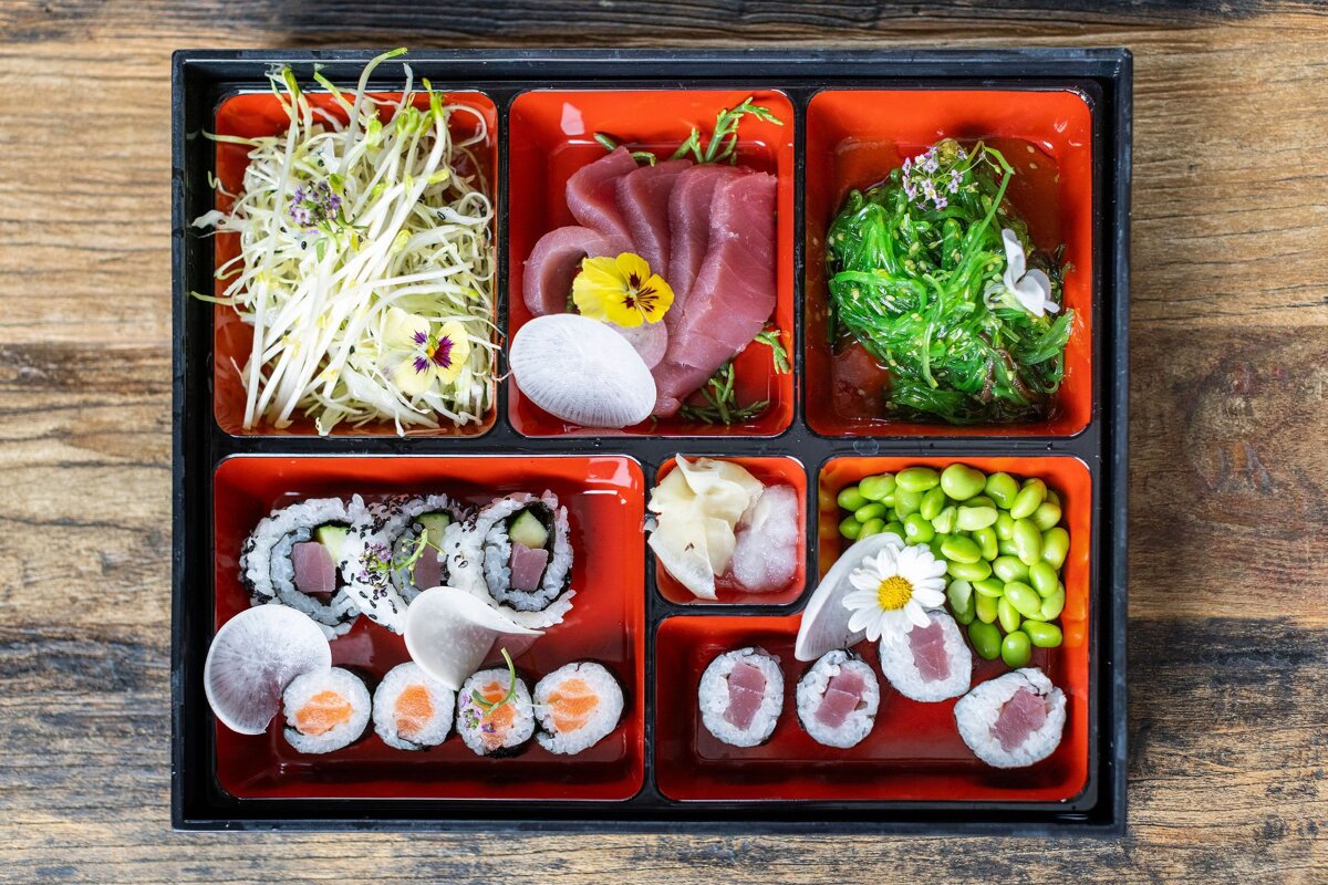 A vibrant Japanese bento box filled with sushi, sashimi, seaweed salad, bean sprouts, edamame, and garnished with flowers on a wooden surface.