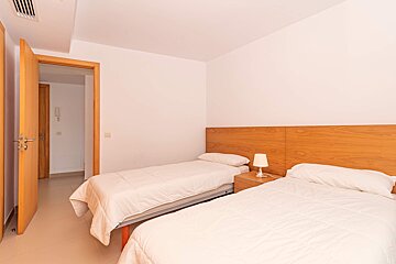 A bright, minimalist bedroom with two twin beds, white bedding, wooden headboards, a bedside lamp, and an open wooden door on the left.