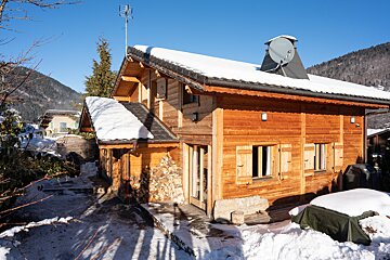 A rustic wooden chalet stands snow-covered on a sunny winter day, with mountains in the background under a clear blue sky.