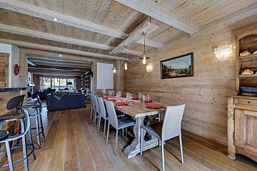 A rustic wooden dining room with a long table, modern chairs, and pendant lights. A cozy living area is visible beyond, creating a warm, inviting chalet interior.