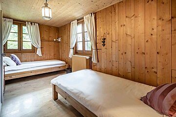 A cozy, rustic bedroom featuring two single beds, wood-paneled walls and ceiling, light wooden floor, and windows with white curtains.
