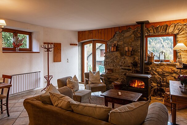 A cozy, rustic living room features a stone wall, a warm wood-burning stove, comfortable sofas, and windows overlooking natural scenery.